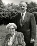 A man standing next to a woman sitting, both posing for this black and white photograph.