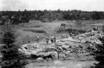Black and white image of dam in the midst of construction. Bolders and pine in foreground. Several men observing and working.
