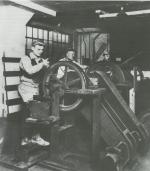 Three men holding large panes of glass against a belt, stand in back of wheels that carry the sharpening belts.'
