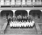 Hershey Industrial School photograph features students posing on steps of Homestead; four rows of boys standing on steps below school sign; front two rows are younger boys in white; back two rows are older boys in dark suits, 1915-1920.