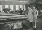 A worker is capping off window glass cylinders preparatory to flattening, while another worker watches.