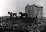 Black and white image of two mules, three students, and the school in the background. 