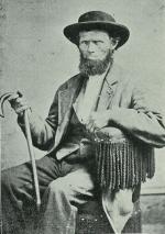 A bearded man wearing a hat and suit, holding a cane, sits in a chair.