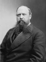 Black and white photograph of a balding, bearded man wearing a suit. 