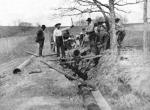 A black and white photograph of group of men dropping a large pipe into a shallow trench using wooden planks as levers.