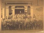 Group photograph of men dressed in suits, most are wearing or holding hats. They are stand in front of the 1868 college building at 145 N Tenth Street.