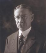 Black and white, head and shoulders image of man with a mustache, wearing spectacles, a suit with a vest and a tie.