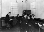 Four male radio broadcasters sit at desk in a staion room.