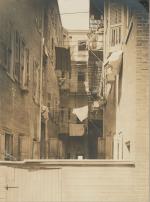 Tenement housing on both sides of a narrow alley. Laundry hangs between the two buildings. In the top right, a mother and children pose on a balcony 