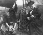 Image of Mother Jones tying a child's shoelace while sitting in a tent. Another woman sits next to her, with a child on her lap and attends to his clothing.
