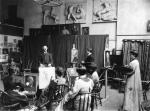 Photograph of an early class with students.   An elderly gentleman poses for students. Five students sit in chairs in front of their easels, while two stand. Art is stored throughout the room.