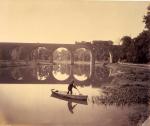 William H. Rau Photograph of Conestoga Bridge, 1891.   A lone man stands in his boat and pushes it along the water with an oar. A train passes over the Conestoga Bridge as the sun casts shadows of the bridge, the train, the man, and his boat. 
