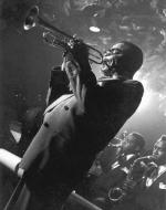 A black and white photo of Dizzy Gillespie playing the trumpet in a club. Visible in the background are members of his band.