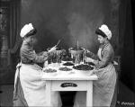 A black and white 1903 image of two Heinz girls, wearing long dresses, white caps and aprons,  filling bottles at studio.