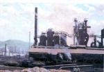 Painting of two steel workers in the foreground and the steel mill with smoke stacks in the background.'