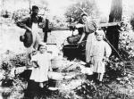 Black and white image of man and woman with two young girls preparing for wash day on the farm. A big black kettle hanging over an open fire holds the water. A wash tub sits on the ground and one can see the wash board placed inside. Buckets for carrying water are in the children's hands.