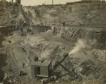 Several miners in ore pit with mining equipment. Photograph shows railroad tracks and steep inclined plane in background used to transport the ore out of the pit.