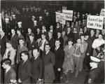Rows of men waiting in lines to enlist.Some carry signs, which read Loose lips sink ships and other slogans.