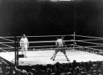 A crowd surrounds a boxing arena as two men fight and a referee looks on.