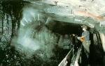 This is color photograph of a longwall mine in operation. The longwall mining machine is working its way along the coalface. A miner wearing a surgical mask and is seen operating the machine. 